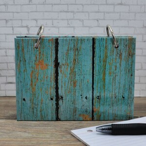 May include: A blue and yellow wood-grain patterned binder with silver rings. The binder is closed and sitting on a wooden surface with a white lined notepad and a black pen.