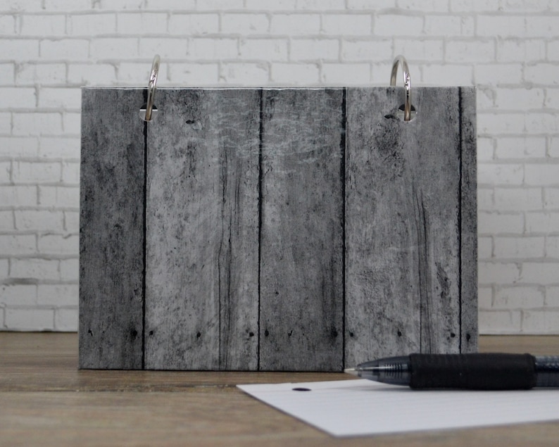 Op de afbeelding: Een grijze binder met houtlook en twee zilveren ringen. De binder staat op een wit bakstenen oppervlak met een zwarte pen ernaast.