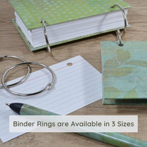 May include: Three silver binder rings of different sizes, a lined index card, a pen, and two small notebooks with patterned covers. The notebooks have silver binder rings. The text "Binder Rings are Available in 3 Sizes" is at the bottom of the image.