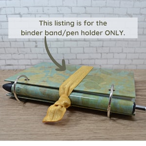 May include: A gold elastic binder band with a pen holder. The band is tied around a blue and green patterned notebook with silver rings.