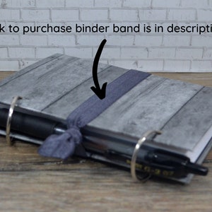 Op de afbeelding: Een grijs en wit notitieboek met een houtnerfpatroon op de omslag. Het notitieboek heeft een zwart elastiek en twee zilveren ringen. Een zwarte pen is in de ringen gestoken.