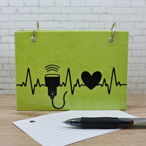 May include: A green notebook with a black and white illustration of a heart beat with a stethoscope and a heart. The notebook has two silver rings on the left and right sides.