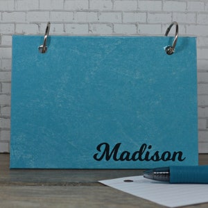 May include: A turquoise blue, textured,  binder with two silver rings. The binder has the name "Madison" printed in black cursive lettering.