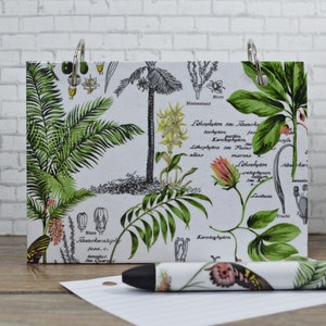 May include: A white notepad with a botanical print featuring green leaves, flowers, and black and white illustrations. The notepad has two silver rings and the text "Lithophyton seu Tzanoko", "Lithophyton seu Fucur", "Lactuca", and "Frucht" is visible.