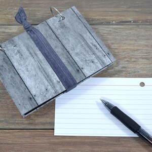 Op de afbeelding: Een grijs en wit notitieboek met een houtnerfpatroon, een zwarte haarband en zilveren ringen. Een witte indexkaart met rode lijnen en een zwarte pen ligt op de tafel naast het notitieboek.