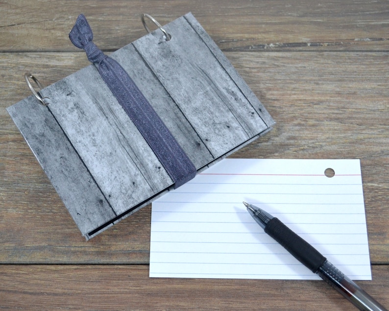 Op de afbeelding: Een grijs en wit notitieboek met een houtnerfpatroon, een zwarte haarband en zilveren ringen. Een witte indexkaart met rode lijnen en een zwarte pen ligt op de tafel naast het notitieboek.