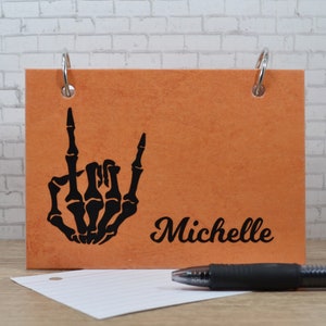 May include: Orange notebook with a black skeleton hand making a rock and roll sign. The notebook has a silver ring binder and the name "Michelle" is printed on the front.