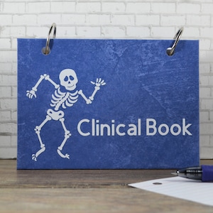 May include: A blue Clinical Book with a white skeleton illustration. The book is held together by two silver rings. A pen and a piece of paper are on the wooden surface in front of the book. The background is a white brick wall.