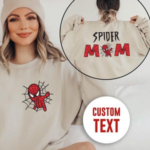 May include: A beige crewneck sweatshirt featuring a red and black spider-themed graphic on the chest. The back design displays the text SPIDER MOM in red block letters with a small spider graphic, and a circular inset shows the back view.