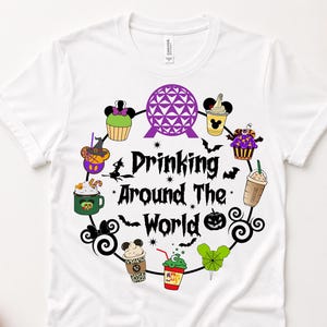 May include: A white short-sleeve t-shirt featuring a circular graphic with the text Drinking Around The World in black. The design includes a purple geodesic dome, various colorful beverage cups, cupcakes, and small black bats and pumpkins.