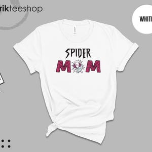 May include: A white short-sleeve t-shirt featuring the black text SPIDER and the word MOM in a dark pink spiderweb pattern. Between the letters, a small white and purple spider-themed character is centered within a web design on the chest.