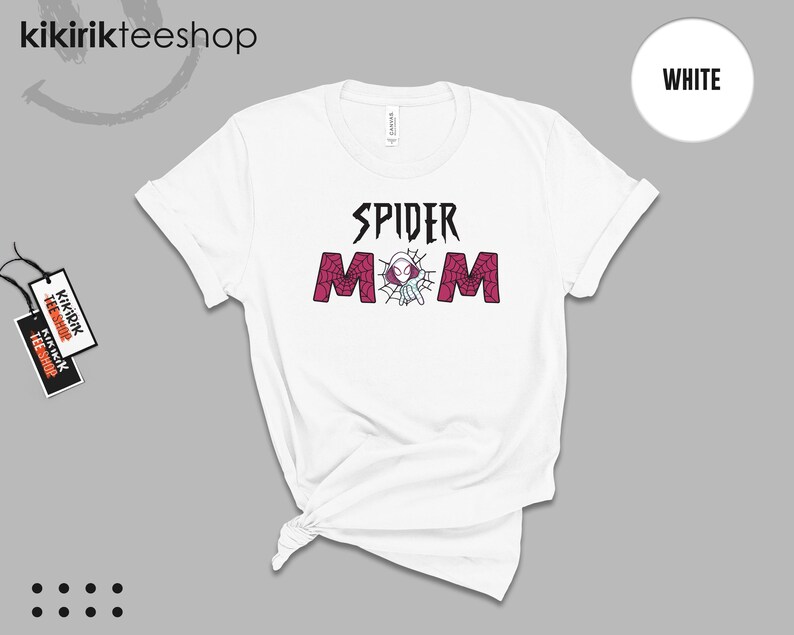 May include: A white short-sleeve t-shirt featuring the black text SPIDER and the word MOM in a dark pink spiderweb pattern. Between the letters, a small white and purple spider-themed character is centered within a web design on the chest.