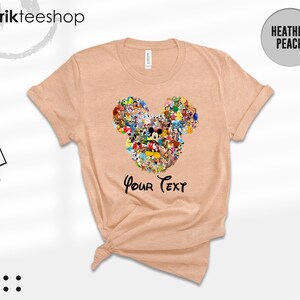 Pu&ograve; includere: T-shirt color pesca con un design a forma di testa di Topolino composto da vari personaggi dei cartoni animati. Il testo "Your Text" &egrave; stampato sotto. La maglietta ha le maniche arrotolate e un'etichetta "Kikirikteeshop".