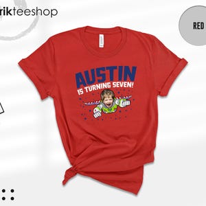 K&ouml;nnte beinhalten: Ein rotes Kurzarm-T-Shirt mit einer Grafik eines Kindes in einem Weltraum-Ranger-Kost&uuml;m mit Fl&uuml;geln. Der Text AUSTIN IS TURNING SEVEN! ist in blauen und wei&szlig;en Buchstaben gedruckt, umgeben von kleinen blauen Sternen. Ein Markenetikett ist befestigt.