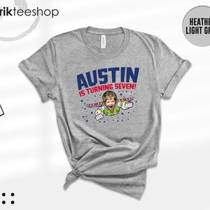 K&ouml;nnte beinhalten: Ein hellgrau meliertes T-Shirt mit einer Grafik, die das Gesicht eines Kindes auf dem K&ouml;rper einer Weltraumfigur mit Fl&uuml;geln zeigt. Der Text AUSTIN IS TURNING SEVEN! steht in blauen und roten Buchstaben, umgeben von kleinen blauen Sternen.