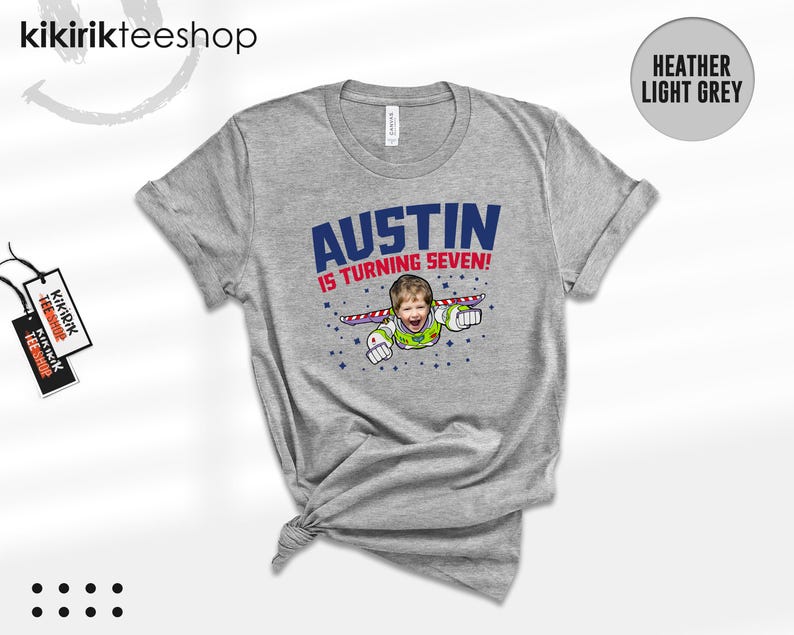 K&ouml;nnte beinhalten: Ein hellgrau meliertes T-Shirt mit einer Grafik, die das Gesicht eines Kindes auf dem K&ouml;rper einer Weltraumfigur mit Fl&uuml;geln zeigt. Der Text AUSTIN IS TURNING SEVEN! steht in blauen und roten Buchstaben, umgeben von kleinen blauen Sternen.