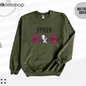 May include: A military green crewneck sweatshirt featuring a graphic print on the chest. The design includes the text SPIDER in black, with the letters S and S filled with a pink spiderweb pattern, and a central illustration of a hooded character.