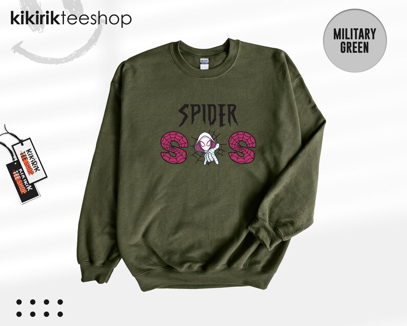 May include: A military green crewneck sweatshirt featuring a graphic print on the chest. The design includes the text SPIDER in black, with the letters S and S filled with a pink spiderweb pattern, and a central illustration of a hooded character.