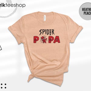 Puede incluir: Camiseta color melocot&oacute;n jaspeado con las palabras "SPIDER PA" en negro y rojo. La palabra "SPIDER" est&aacute; en una fuente estilizada, y "PA" est&aacute; en negrita, fuente roja con un dise&ntilde;o de telara&ntilde;a. La camiseta tiene mangas cortas y cuello redondo.