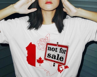May include: White t-shirt with a red and white outline of Canada and a red maple leaf. A red and white sign with the text "not for sale" is placed over the outline of Canada.