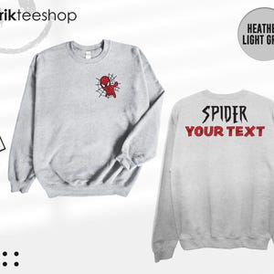 May include: A heather light grey crewneck sweatshirt featuring a small red and black spider-themed character graphic on the front left chest. The back of the sweatshirt displays the black text SPIDER and the red text YOUR TEXT in a bold font.