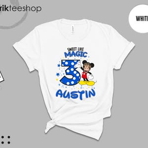 May include: A white short-sleeve t-shirt featuring a graphic print with the text SWEET LIKE MAGIC, the number 3 in blue with white polka dots, and the name AUSTIN. A cartoon character with a child's face is positioned next to the number 3.