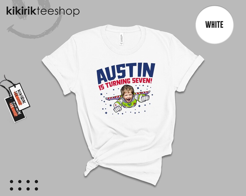 K&ouml;nnte beinhalten: Ein wei&szlig;es T-Shirt mit einem Grafikdruck, der das Gesicht eines Kindes auf dem K&ouml;rper einer Weltraumfigur mit Fl&uuml;geln zeigt. Der Text AUSTIN IS TURNING SEVEN! ist in blauen und roten Buchstaben gedruckt, umgeben von kleinen blauen Sternsymbolen.