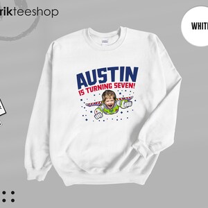 K&ouml;nnte beinhalten: Ein wei&szlig;es Sweatshirt mit Rundhalsausschnitt und einem Grafikdruck, der das Gesicht eines Kindes auf dem K&ouml;rper einer Weltraum-Ranger-Figur zeigt. Der Text AUSTIN IS TURNING SEVEN! steht in blauen und roten Buchstaben, umgeben von kleinen blauen Sternen.