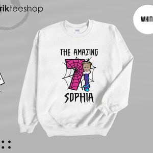 May include: A white crewneck sweatshirt featuring a graphic print of a large pink number seven with a black spiderweb pattern. The text THE AMAZING SOPHIA is printed in black above and below the number, with a small photo of a child's face.