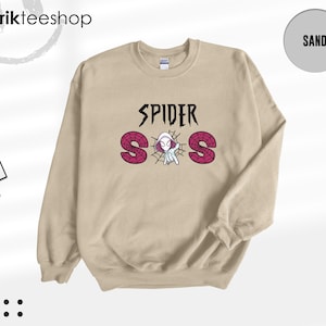 May include: A sand-colored crewneck sweatshirt featuring a graphic print on the chest. The text reads SPIDER SOS in black and pink, with a small white and pink spider-themed character positioned in the center of a black spiderweb design.