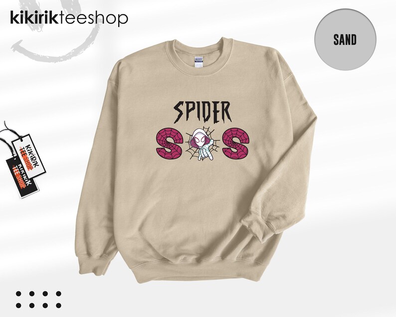 May include: A sand-colored crewneck sweatshirt featuring a graphic print on the chest. The text reads SPIDER SOS in black and pink, with a small white and pink spider-themed character positioned in the center of a black spiderweb design.