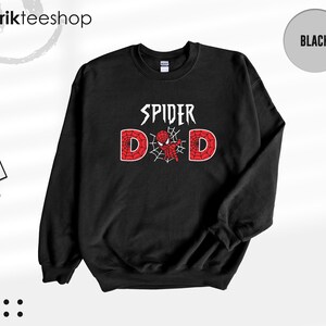 May include: A black long-sleeve crewneck sweatshirt featuring the white text SPIDER above the word DAD, where the letter A is replaced by a red and black spider-themed graphic with a web design. The sweatshirt has a ribbed collar and cuffs.