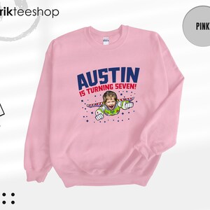 K&ouml;nnte beinhalten: Ein hellrosa Rundhals-Sweatshirt mit einem Grafikdruck, der den Text AUSTIN IS TURNING SEVEN! &uuml;ber einer Illustration eines Kindergesichts auf einem Weltraum-Charakterk&ouml;rper mit Fl&uuml;geln zeigt, umgeben von kleinen dunkelblauen Sternen.