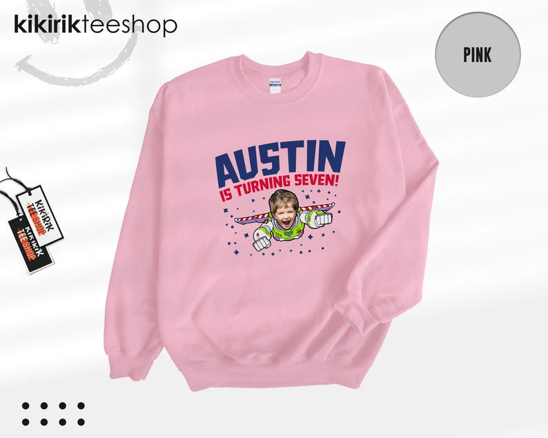 K&ouml;nnte beinhalten: Ein hellrosa Rundhals-Sweatshirt mit einem Grafikdruck, der den Text AUSTIN IS TURNING SEVEN! &uuml;ber einer Illustration eines Kindergesichts auf einem Weltraum-Charakterk&ouml;rper mit Fl&uuml;geln zeigt, umgeben von kleinen dunkelblauen Sternen.
