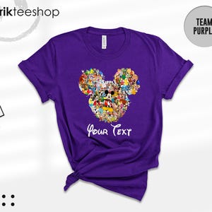 Pu&ograve; includere: Una maglietta viola con un design a forma di testa di Topolino composto da vari personaggi dei cartoni animati. La maglietta ha le maniche arrotolate e la scritta "Your Text" in bianco. Il marchio "kikirikteeshop" &egrave; visibile in alto.