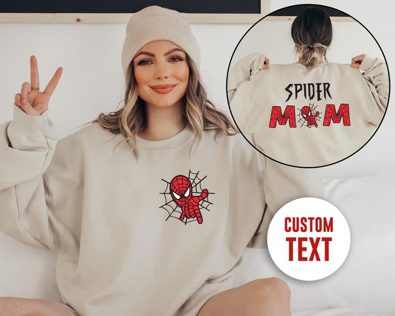 May include: A beige crewneck sweatshirt featuring a red and black spider-themed graphic on the chest. The back design displays the text SPIDER MOM in red block letters with a small spider graphic, and a circular inset shows the back view.