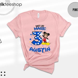 May include: A light pink t-shirt featuring a graphic with the text SWEET LIKE MAGIC, the number 3 in blue with white polka dots, and the name AUSTIN. A cartoon character with a child's face is positioned next to the number 3.