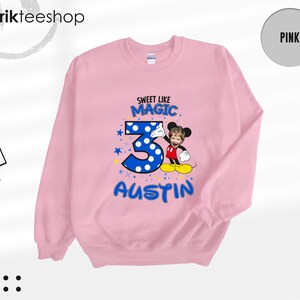 May include: A light pink long-sleeve sweatshirt featuring a graphic print with the text "SWEET LIKE MAGIC", a large blue polka-dot number "3", and the name "AUSTIN" in blue. A cartoon character with a child's face is positioned next to the number.