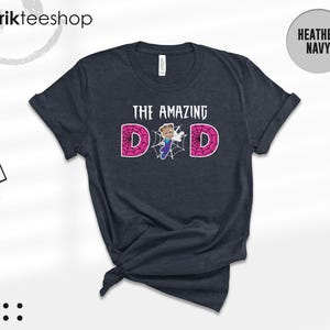 May include: A heather navy short-sleeve t-shirt featuring the text THE AMAZING DAD in white, with the letter A replaced by a graphic of a person in a blue and purple suit and the letters D and D filled with a pink spiderweb pattern.