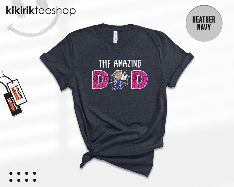 May include: A heather navy short-sleeve t-shirt featuring the text THE AMAZING DAD in white, with the letter A replaced by a graphic of a person in a blue and purple suit and the letters D and D filled with a pink spiderweb pattern.