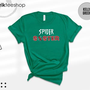 Puede incluir: Camiseta verde Kelly con las palabras "SPIDER SISTER" en blanco y rojo, con un gr&aacute;fico de ara&ntilde;a. La camiseta est&aacute; doblada en la parte inferior. La marca "kikirikteeshop" est&aacute; en la parte superior izquierda.
