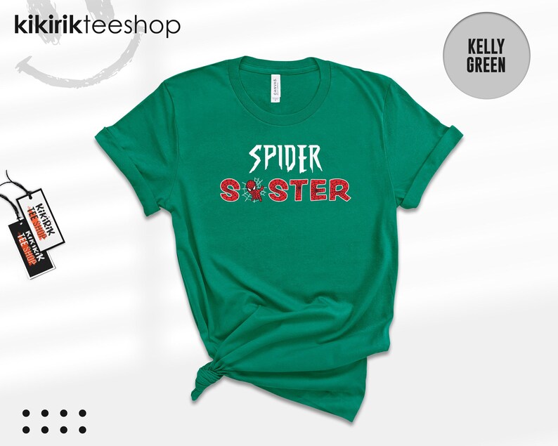 Puede incluir: Camiseta verde Kelly con las palabras "SPIDER SISTER" en blanco y rojo, con un gr&aacute;fico de ara&ntilde;a. La camiseta est&aacute; doblada en la parte inferior. La marca "kikirikteeshop" est&aacute; en la parte superior izquierda.