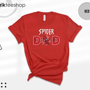 Puede incluir: Una camiseta roja con las palabras "SPIDER DAD" en blanco. La letra "D" est&aacute; dise&ntilde;ada con un patr&oacute;n de telara&ntilde;a y un gr&aacute;fico de Spider-Man. La camiseta tiene cuello redondo y mangas cortas.