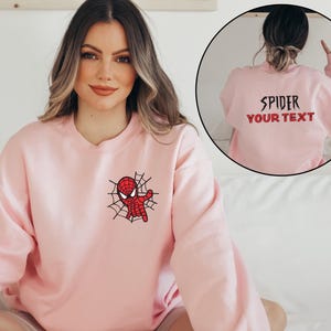 May include: A light pink crewneck sweatshirt featuring a small embroidered graphic of a red and black spider character on a web on the left chest. The back of the sweatshirt displays the black text SPIDER and red text YOUR TEXT.