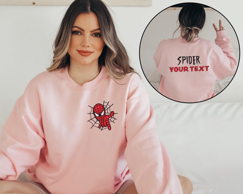May include: A light pink crewneck sweatshirt featuring a small embroidered graphic of a red and black spider character on a web on the left chest. The back of the sweatshirt displays the black text SPIDER and red text YOUR TEXT.