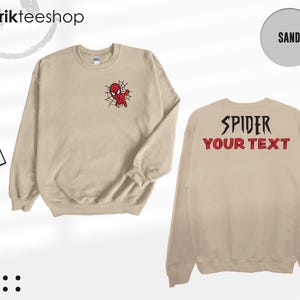 May include: A sand-colored crewneck sweatshirt featuring a small embroidered red and blue spider-themed character on the left chest. The back displays the black text SPIDER and the red text YOUR TEXT in a bold, stylized font.