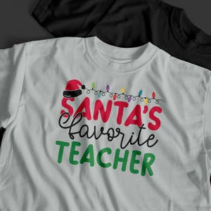 May include: White t-shirt with the text "SANTA'S favorite TEACHER" in red, black, and green. The design includes a Santa hat and string lights. A black t-shirt is partially visible in the background.