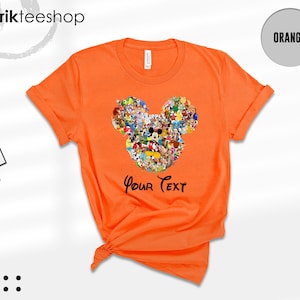 Pu&ograve; includere: T-shirt arancione brillante con un design a forma di testa di Topolino composto da personaggi dei cartoni animati. Il testo "Your Text" &egrave; stampato sotto. La maglietta ha un girocollo e maniche arrotolate.