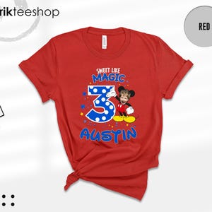 May include: A red short-sleeved t-shirt featuring a graphic print with the text "SWEET LIKE MAGIC" above a large blue polka-dot number "3" and the name "AUSTIN" below. A cartoon character with a child's face is positioned next to the number.