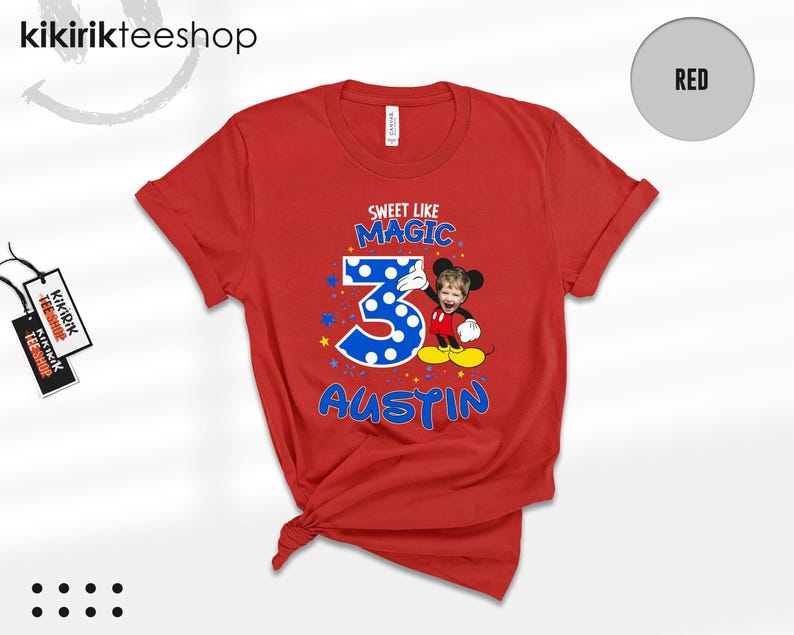 May include: A red short-sleeved t-shirt featuring a graphic print with the text "SWEET LIKE MAGIC" above a large blue polka-dot number "3" and the name "AUSTIN" below. A cartoon character with a child's face is positioned next to the number.